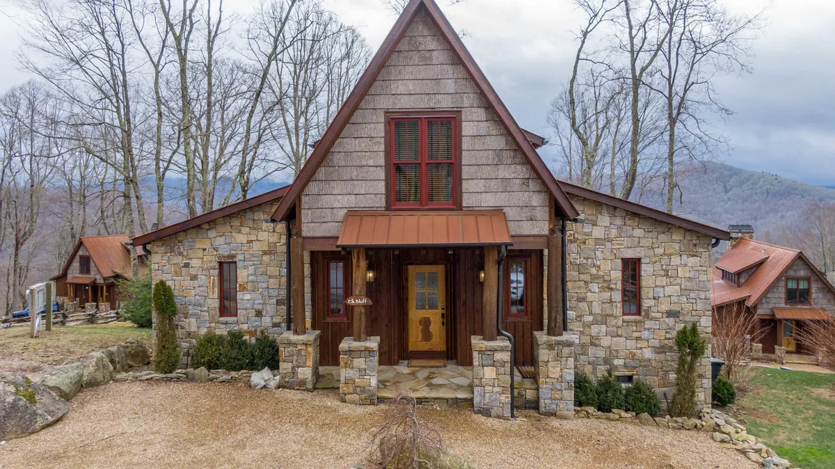 This image features the stone facade and wooden door of Elk Bluff's exterior, nestled among trees with a triangular roof. The setting exudes a welcoming, nature-connected atmosphere, and the visible porch hints at spaces where guests can enjoy mountain air and contemplate the outdoors.