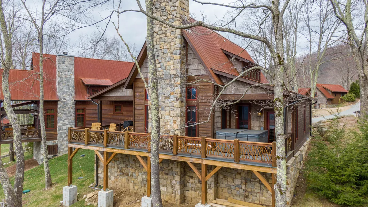A cozy exterior scene of a wooden deck, featuring a hot tub, stone chimney, red metal roof, and comfortable seating. The setting is perfect for group gatherings, sunset watching, and enjoying the outdoors in all seasons.
