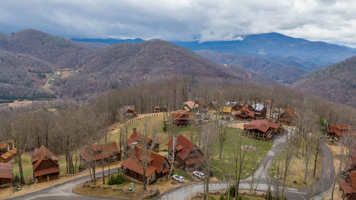A panoramic view of rustic cabins nestled along a roadway, backed by a dramatic mountain range and lush tree line. The scene evokes the natural beauty of the Eagles Nest community, with space for hiking, gathering, and enjoying seasonal changes like fall foliage and morning mist.