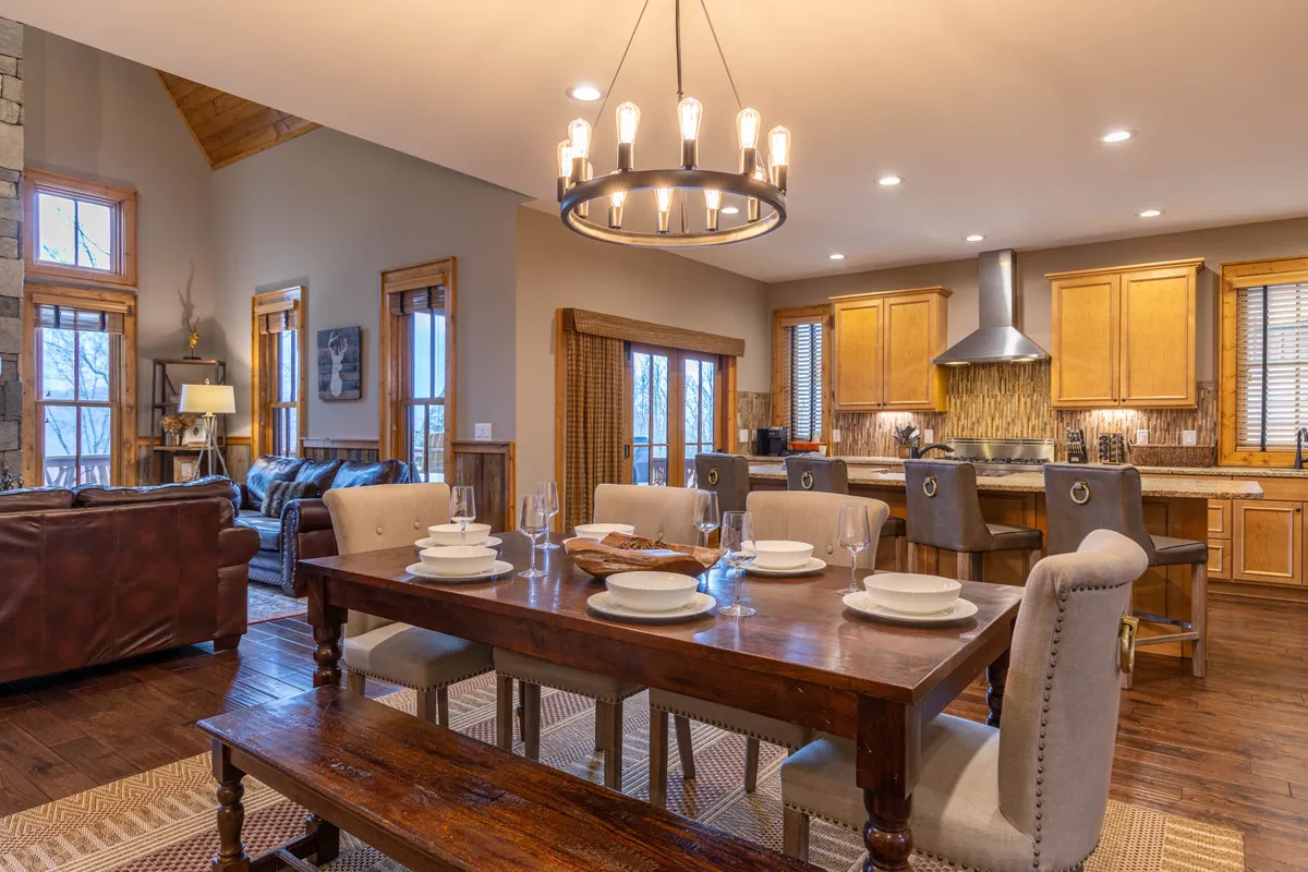 An open-concept dining and kitchen area with a dining table seating six, an elegant chandelier overhead, kitchen appliances and cabinetry in the background, and a cozy seating area visible. The space exudes warmth and encourages togetherness for meals and gatherings.