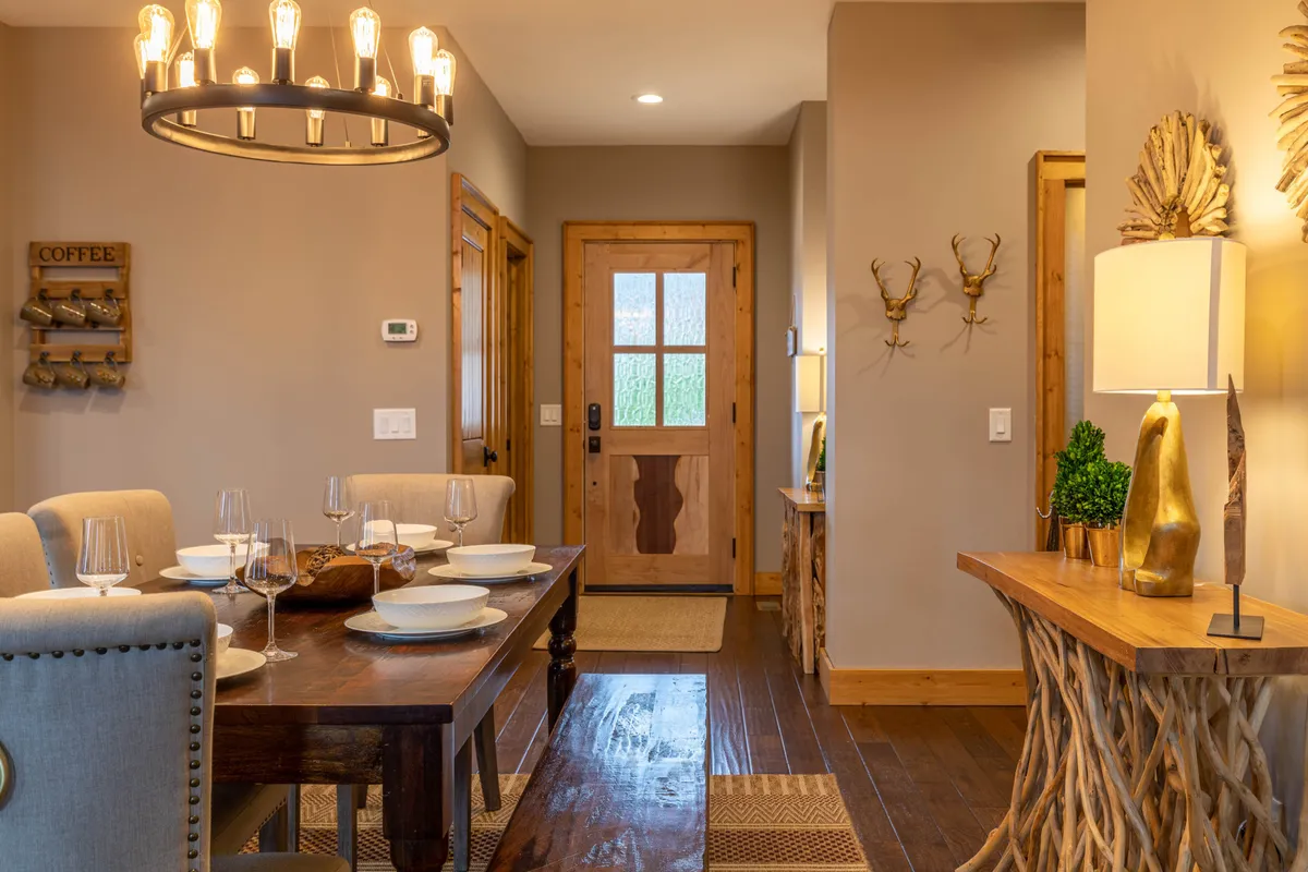 A cozy dining room scene featuring a central table under a warm chandelier, with inviting decor including a side table and decorative antlers. The space is staged for a meal or gathering, evoking warmth and togetherness, suitable for both casual and special occasion dining.