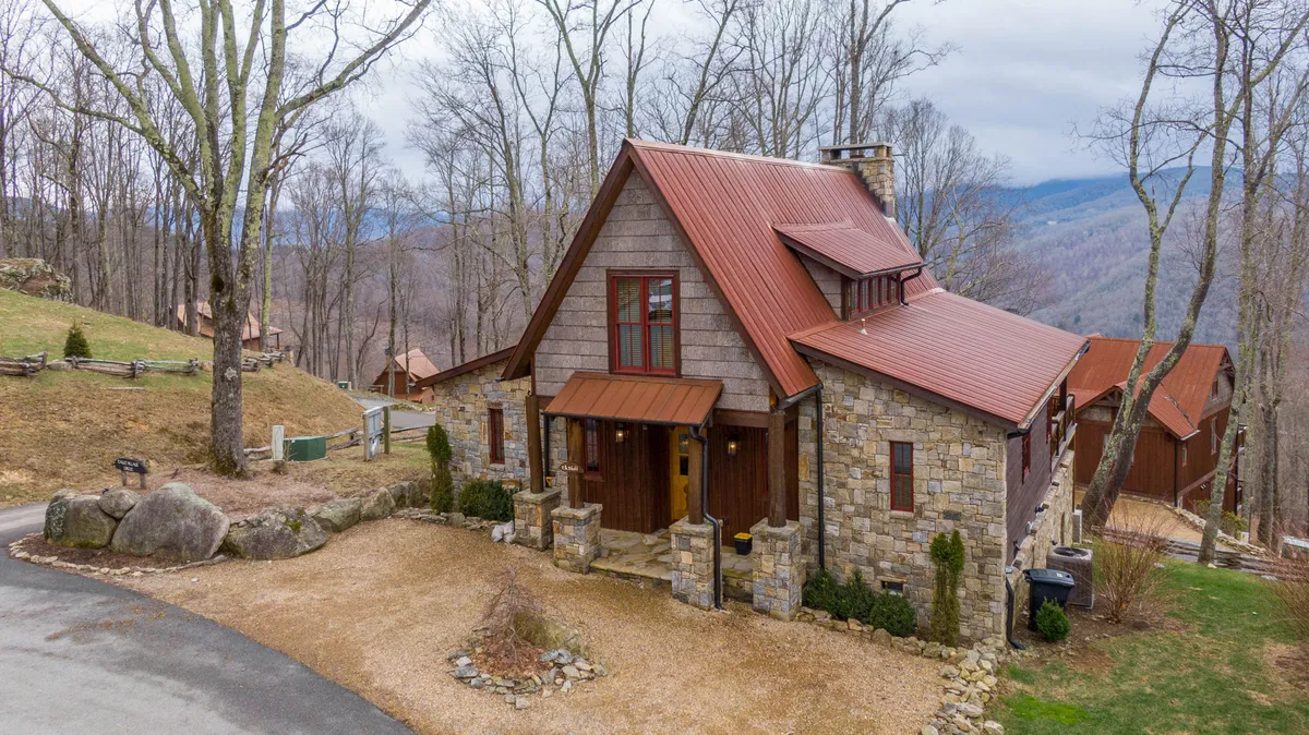The hero image showcases the home's striking stone facade, a classic metal roof, and a welcoming wooden door, all framed by a meticulously landscaped garden. The setting exudes peace and charm, with the inviting porch offering a perfect spot to relax and enjoy sweeping mountain views.