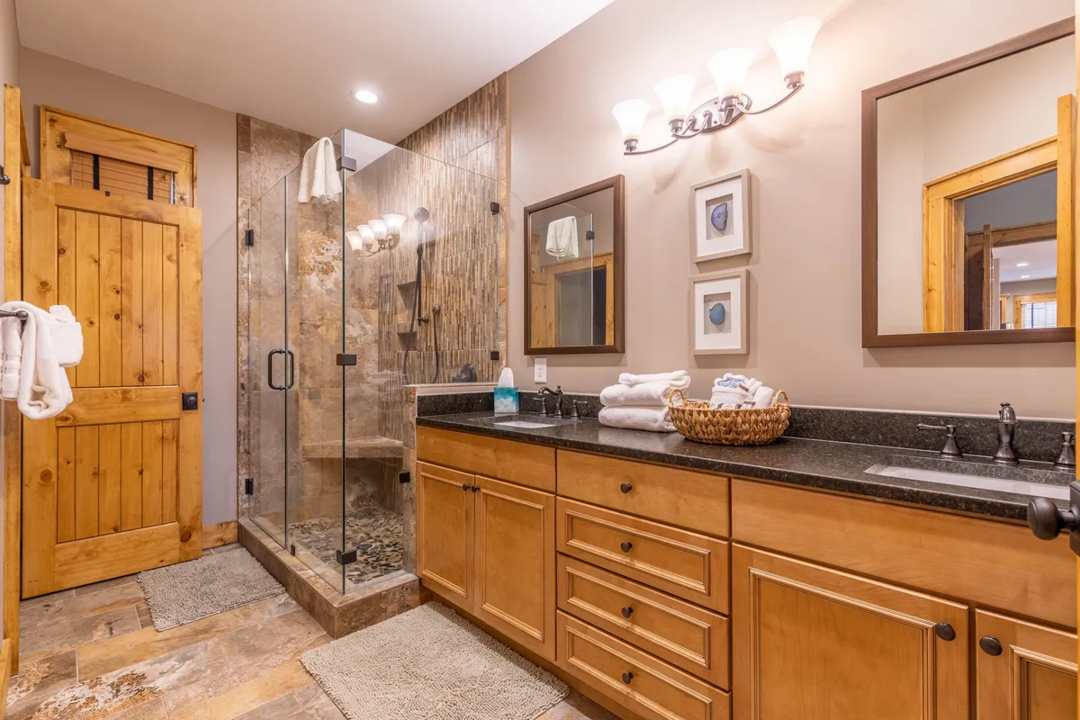 A luxurious ensuite bathroom with a sleek glass shower enclosure, double granite vanities, framed mirrors, and warm wooden accents. The space feels both functional and indulgent, ideal for a relaxing start or end to the day.