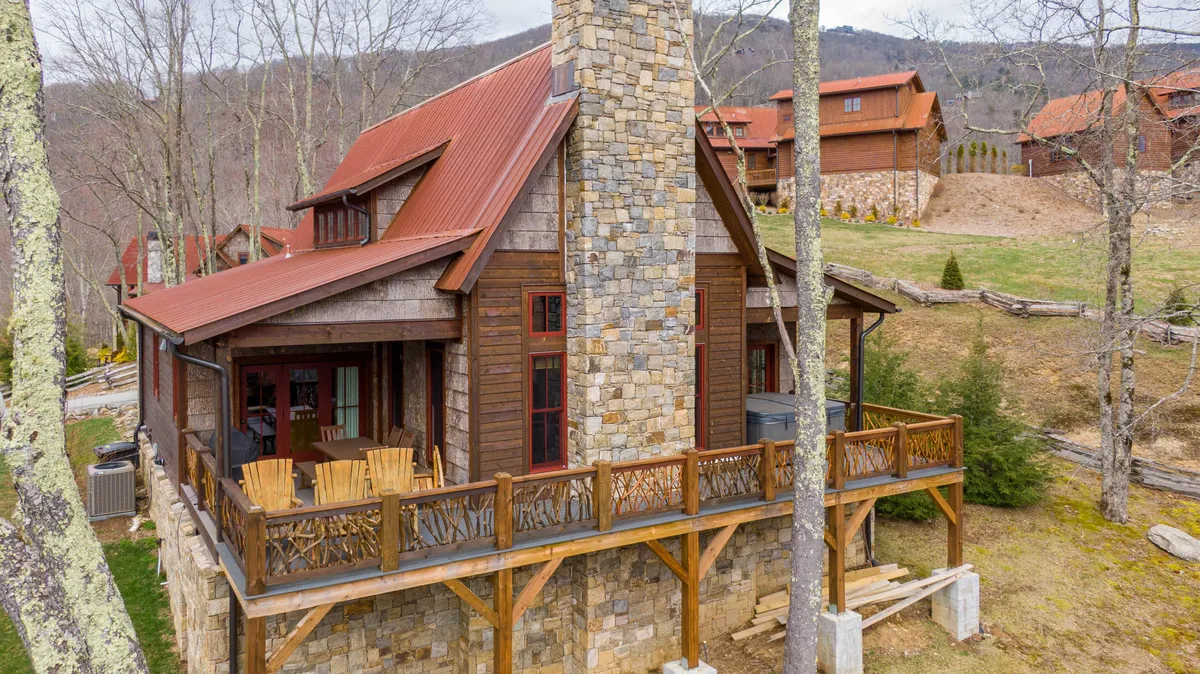 This image features the exterior of a mountain lodge with a stone chimney, wooden balcony, and red roof nestled amid lush trees. The scene highlights an inviting balcony—ideal for relaxing outdoors—and hints at stunning sunset views and stargazing.