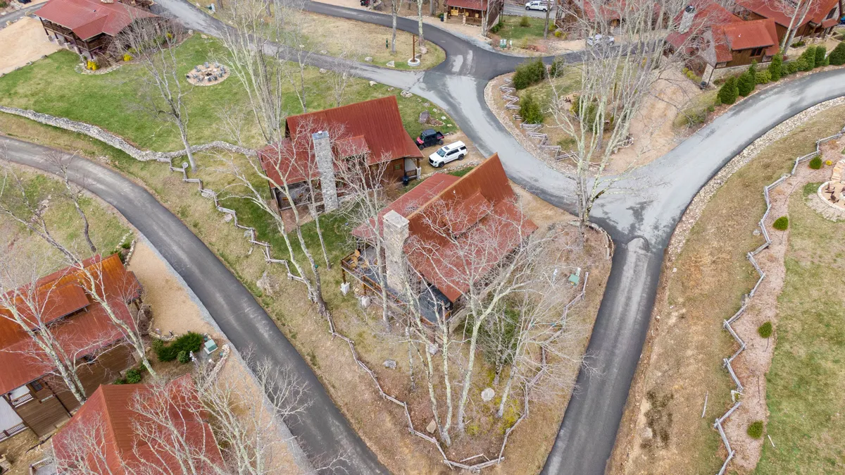 The image shows rustic cabins surrounded by trees, a circular stone fire pit, and winding roads leading through the landscape. It evokes a sense of community while maintaining a peaceful, secluded atmosphere perfect for group gatherings and quiet evenings.