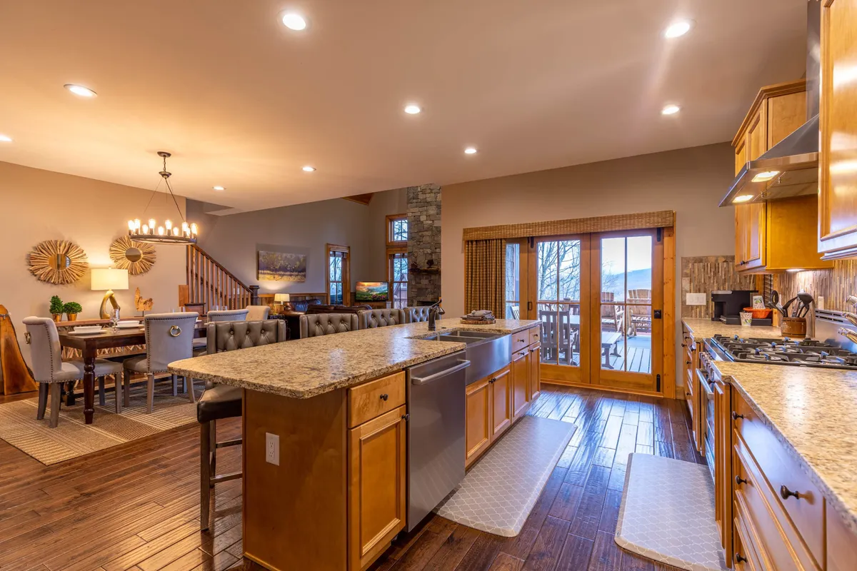 This image captures a warm, active kitchen and dining area with an island countertop, adjacent dining space, and a visible outdoor deck. People are engaged in cooking, entertaining, and gathering, providing a lively and communal atmosphere. The view extends to the outdoors, hinting at the connection between indoor living and exterior amenities.