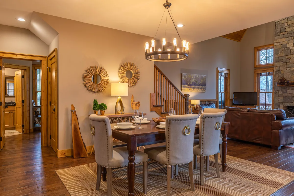 An elegantly set dining table with a chandelier overhead, decorative mirrors on the wall, and a wooden staircase in the background. The room radiates comfort and a touch of formality, ideal for both holiday feasts and informal game nights.