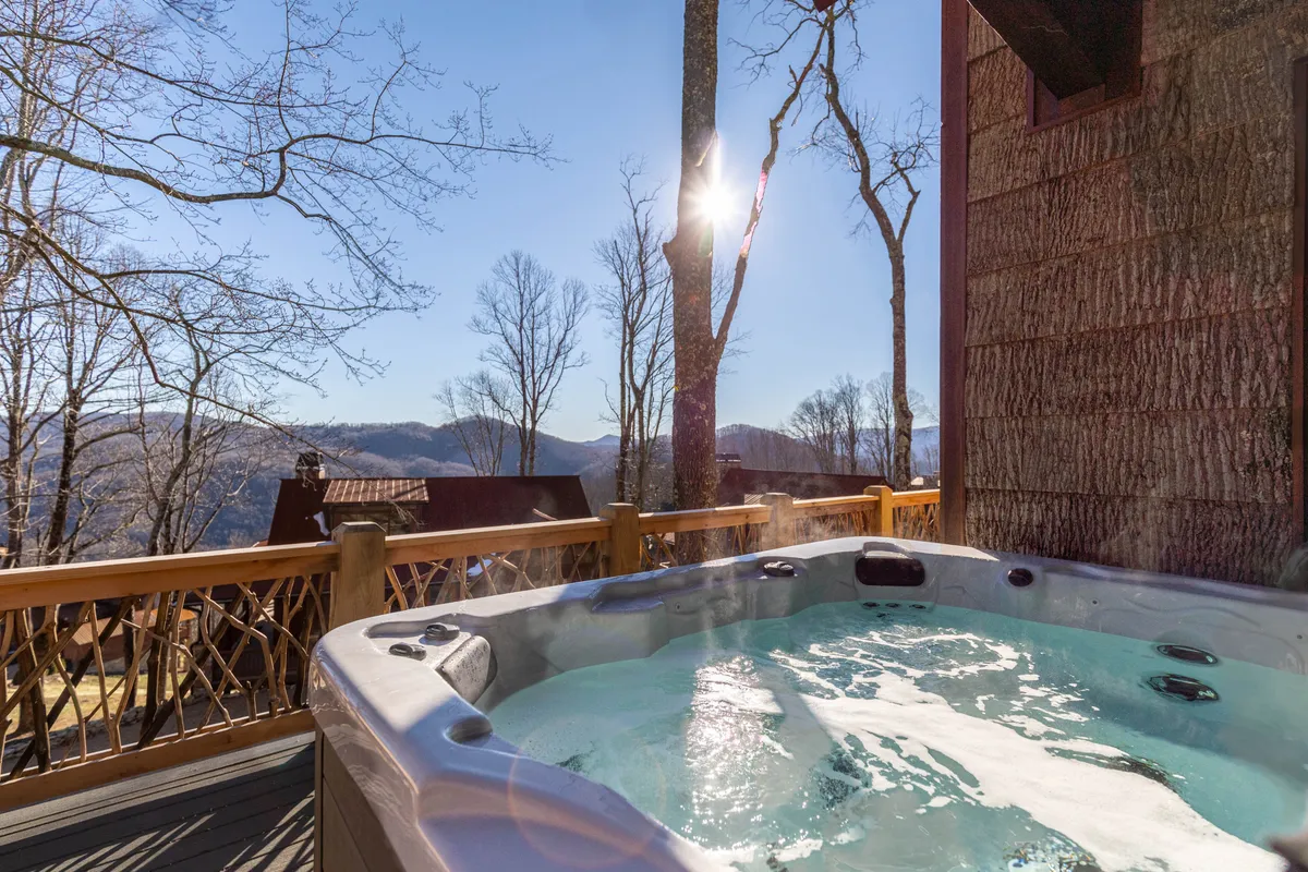 The photo captures a hot tub nestled outdoors, offering a sweeping mountain view framed by sunlight filtering through the trees. The mood is serene, suggesting both daytime relaxation and starlit evenings in the tub.